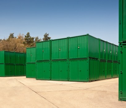 A large outdoor storage area featuring multiple green metal shipping containers stacked two high, situated on a paved surface within a commercial or industrial setting. The containers are rectangular, with ribbed sides and smooth doors, all painted in a uniform green colour with visible locking mechanisms. The background shows a clear blue sky and a line of trees with some sparse foliage, indicating a rural or semi-rural location near Enfield. The containers occupy most of the image, arranged in a neat, organized formation, with some positioned directly on the ground and others stacked above. The scene appears well-lit with natural daylight, emphasizing the clean, freshly painted appearance of the containers. The setting suggests a site used for storing or temporary holding of waste or materials, aligned with services provided by Business Waste Removal Enfield specializing in rubbish management and waste disposal for local clients in the area.